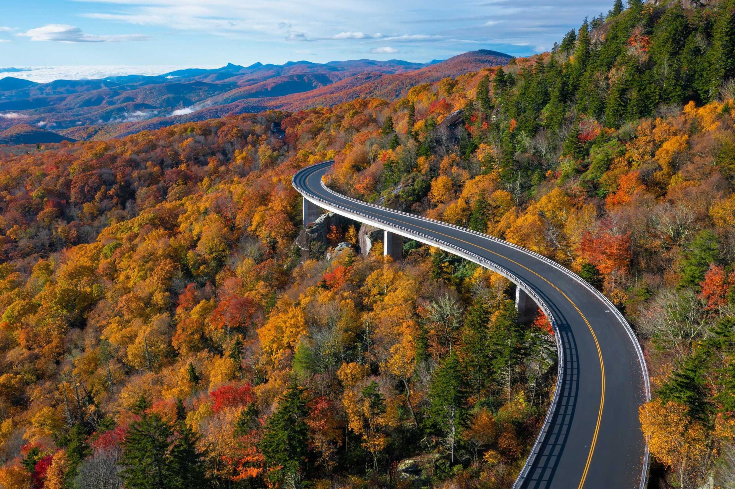 A curving elevated road passes through a forest of autumn-colored trees with mountains visible in the distance on the Blue Ridge Parkway.
