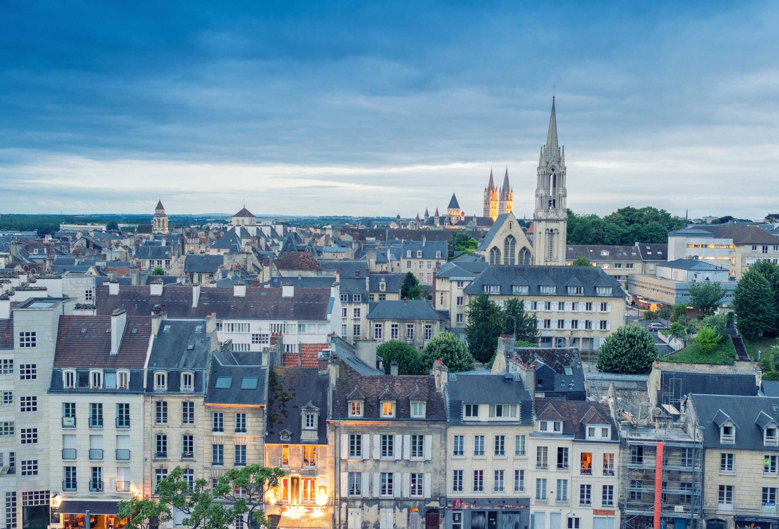 Aerial cityscape of Caen in Normandy at dusk with buildings with grey roofs and a tall spired church.