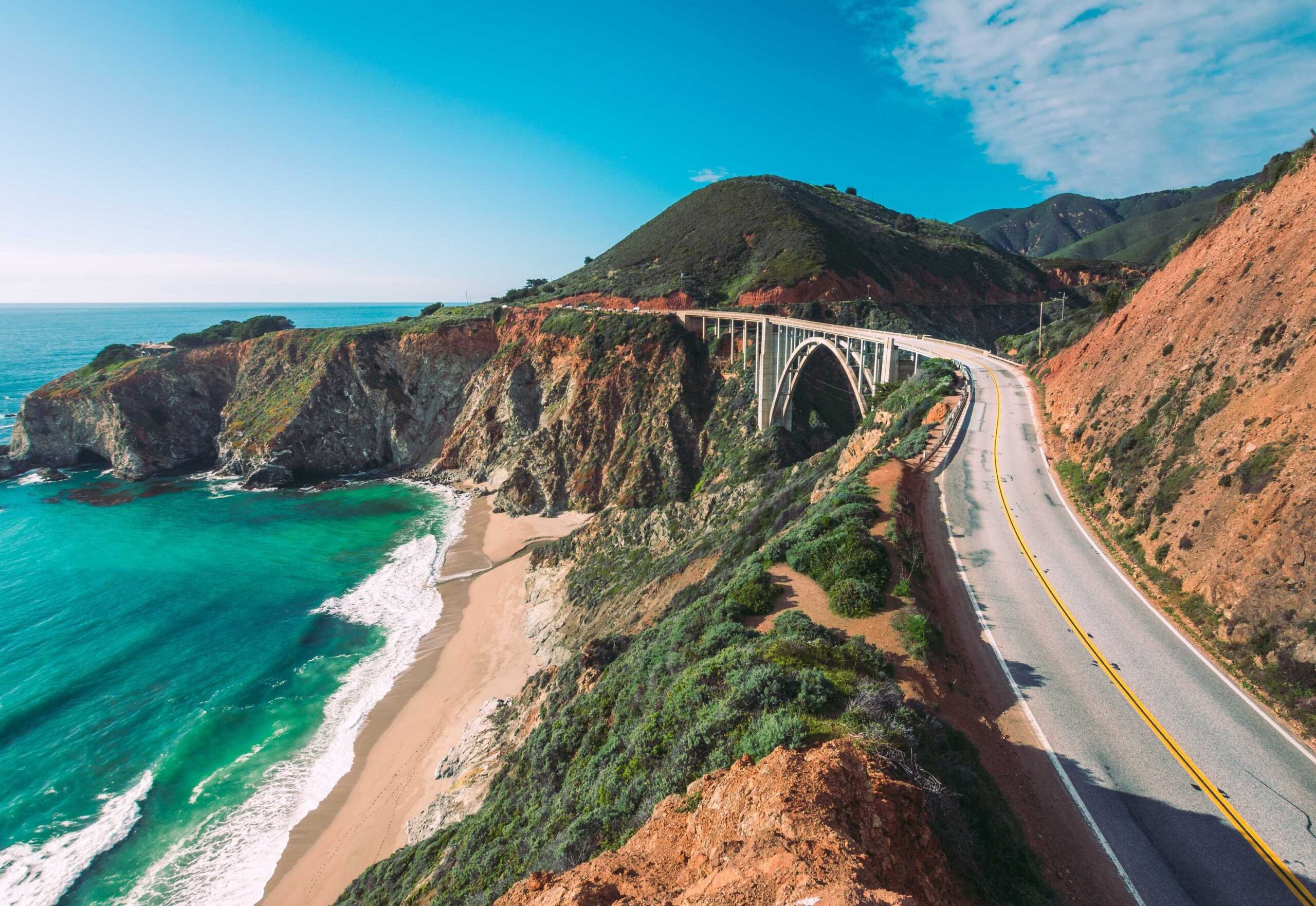 View of a winding road and bridge above cliffs on the Pacific Coast Highway with turquoise ocean waves and a sandy beach below.