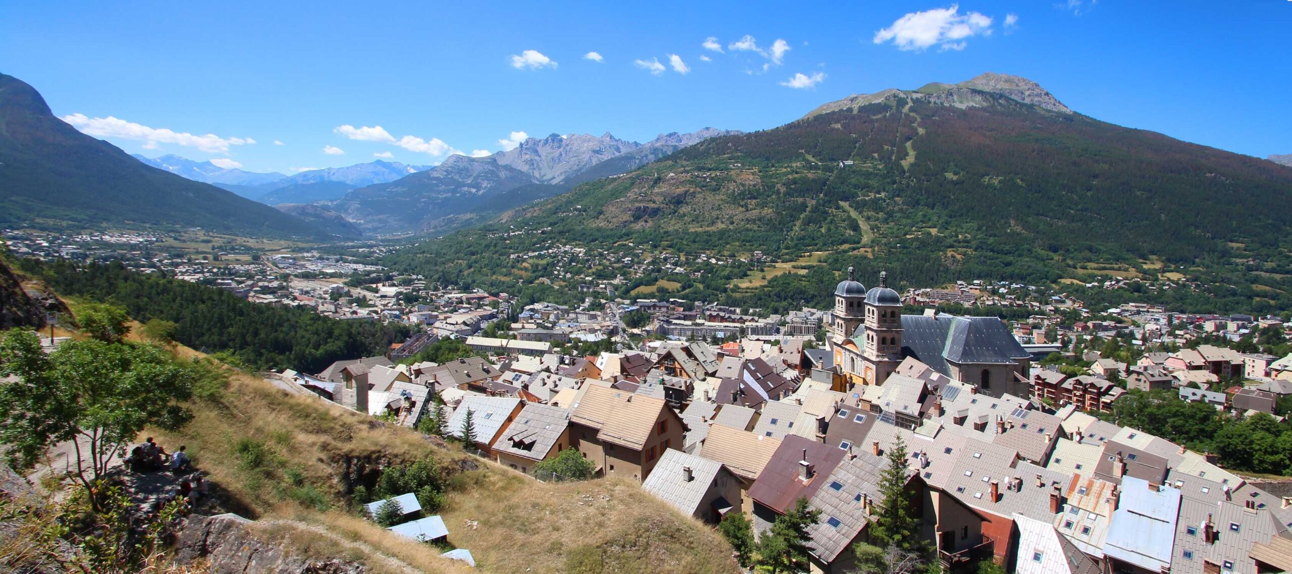View of Briançon with clustered rooftops framed by lush green mountains under a  blue sky.