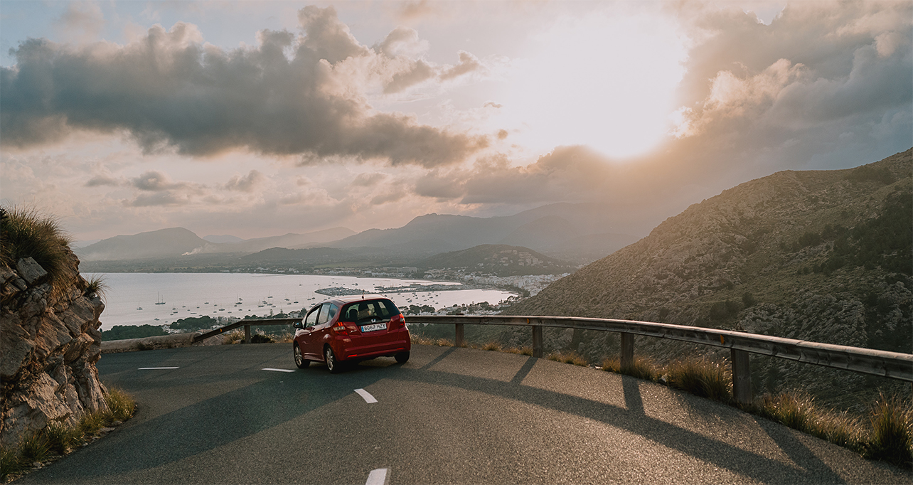 A red car drives along a winding mountain road under a partially cloudy sky at sunset overlooking a serene bay.