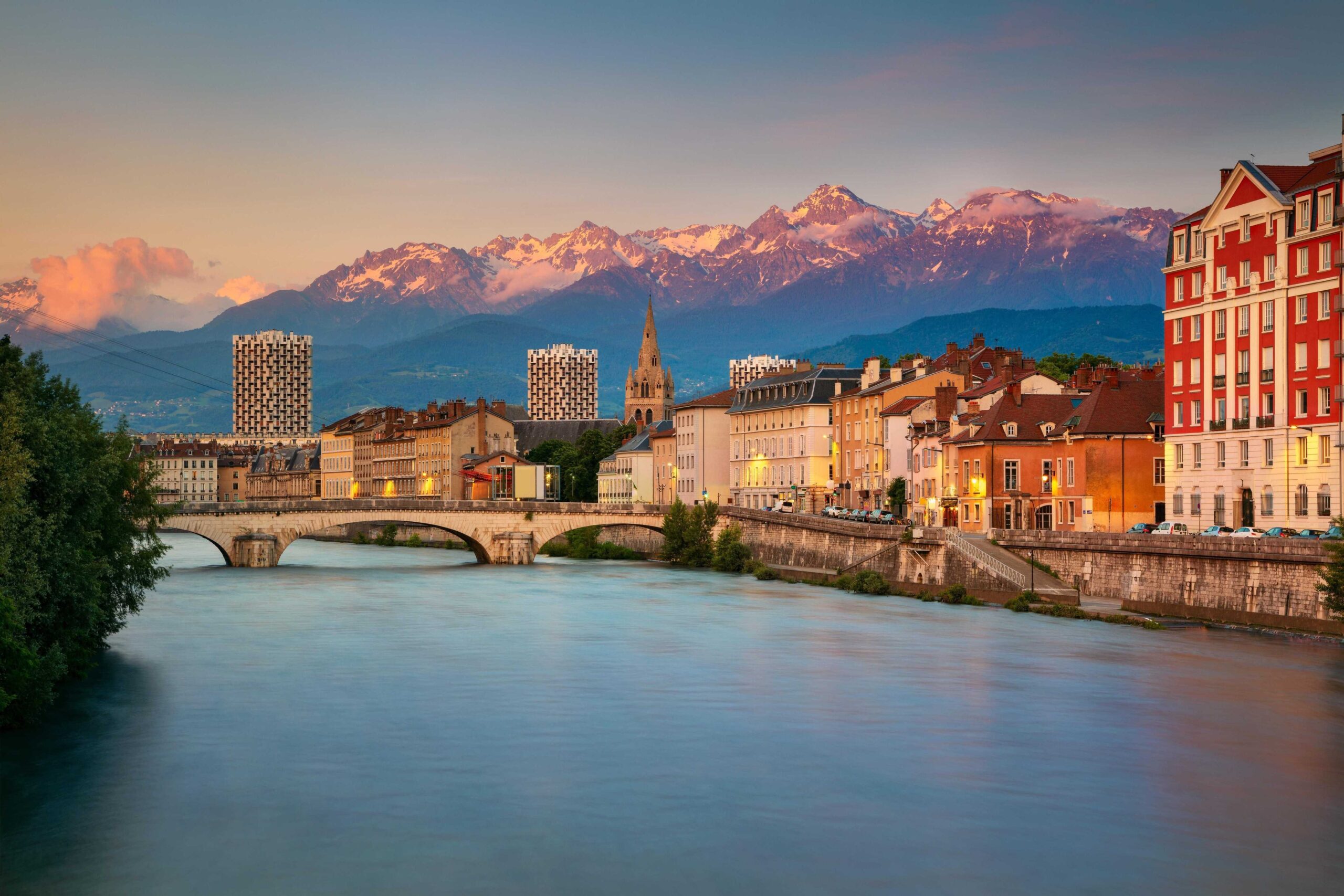 View of Briançon with clustered rooftops framed by lush green mountains under a  blue sky.