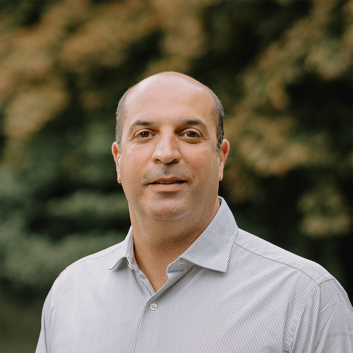 A man smiling in a light gray shirt is standing outdoors with a blurred background of green foliage.