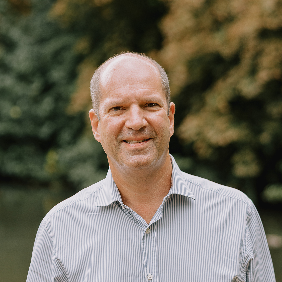 Smiling middle-aged man wearing a striped shirt poses outdoors with soft-focus green foliage in the background.